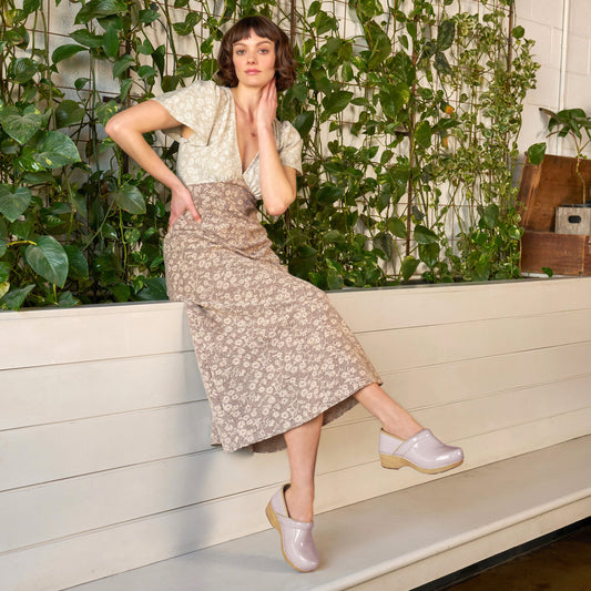 Woman in a floral skirt and light shoes leaning against a white wall with greenery.