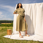 Woman in field standing in front of a neutral fabric backdrop wearing a khaki green outfit and cream wooden mules.
