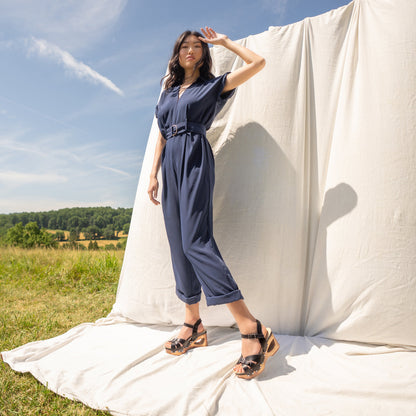 Woman in field in front of neutral backdrop wearing a jumpsuit and black mary jane heels with wood bottoms.