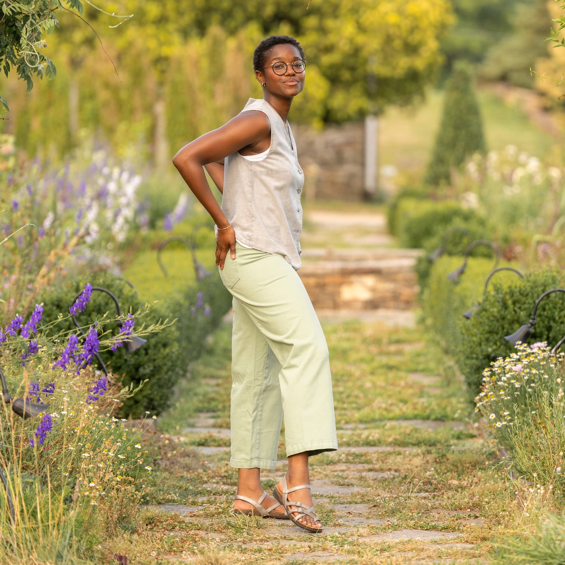 Woman in outdoor garden wearing neutral outfit with metallic strappy flat sandals.